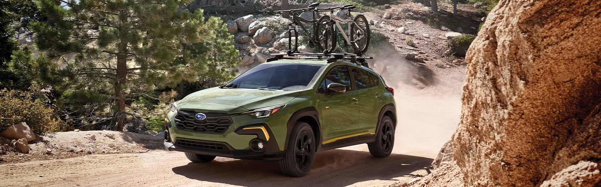 A green Subaru Crosstrek with two bicycles on its roof rack drives on a dusty dirt road through pine trees.