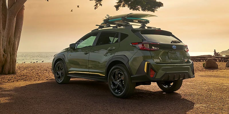 Green Subaru Crosstrek with surfboards on its roof rack, parked overlooking a beach at sunset.