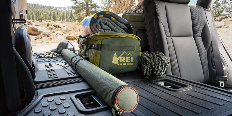 The back of a vehicle is loaded with camping gear, including an REI bag, blanket, fishing rod, and rope.