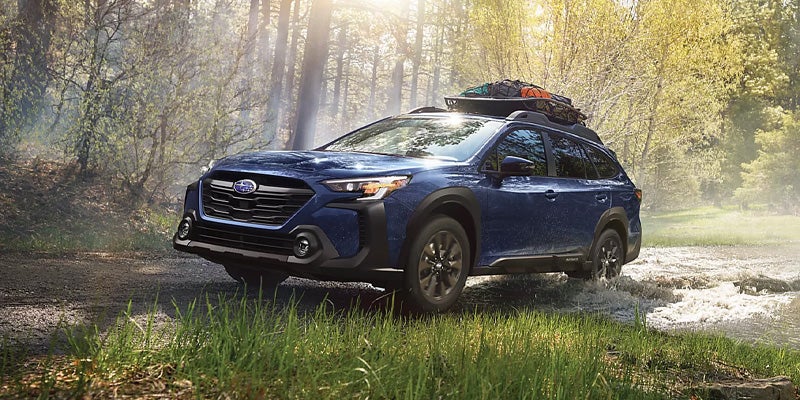 A dark blue Subaru Outback with a roof-mounted cargo carrier splashes through a shallow creek in a sunlit forest.