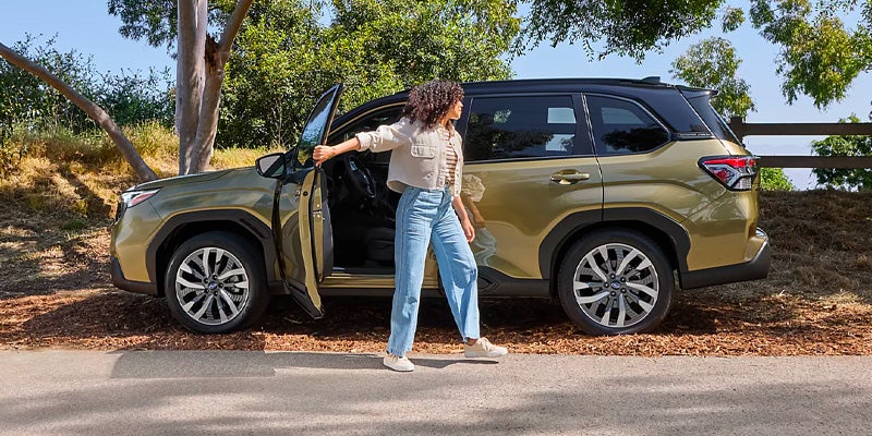 A woman with curly brown hair in a striped shirt, jeans, and cream jacket leans against a green Subaru with the driver's door open, looking right.