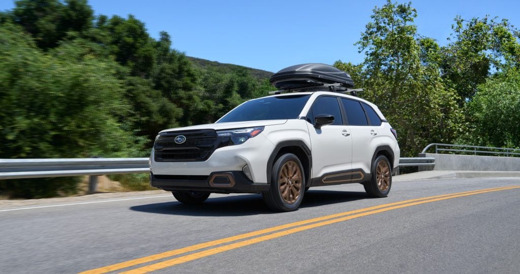 White Subaru SUV driving on a scenic mountain road with a roof cargo carrier.