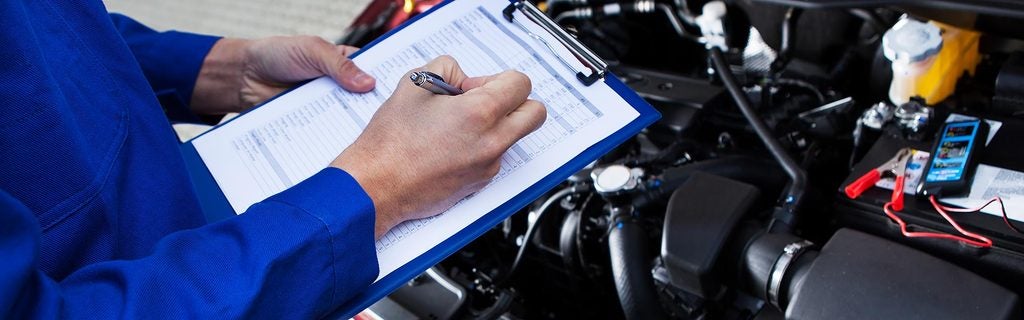Automotive technician completing a vehicle inspection checklist near an open engine bay.